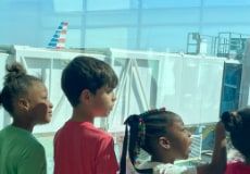Four children stand in a line at a large airport terminal window, looking out at a jet bridge and an aircraft tail.
