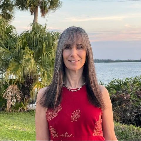 Allison Draper, with long gray hair and a red sleeveless top, standing outdoors by palm trees and water.