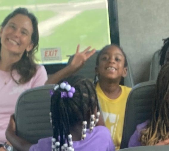 A smiling woman and several children seated on a bus, some waving at the camera.