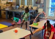 Children play in a classroom with tables, floor mats and a hula hoop.