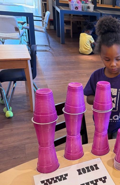 Children at a classroom table stacking pink plastic cups into towers.
