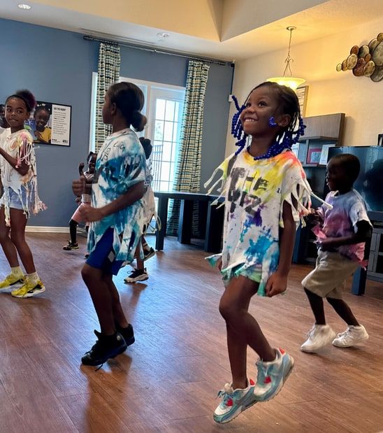 Children in tie-dyed, fringed T-shirts dance across a wood-floored room; a girl with blue beaded braids smiles in the center.