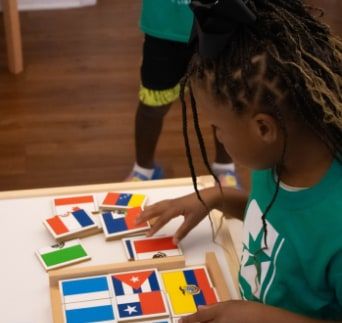 A girl with long braids and a bow, in a green T-shirt, arranges wooden flag puzzle pieces on a table.