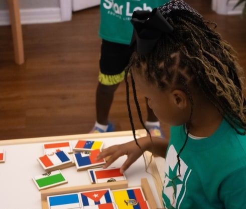 A girl with long braids arranges flag picture tiles on a wooden tray at a table.