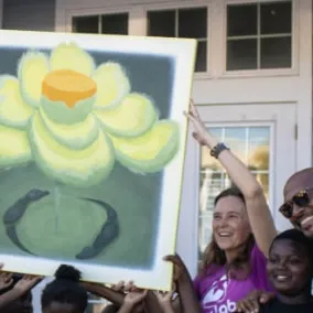 A smiling woman in a purple Star Lab shirt and a man in a suit hold up a large painting of a yellow flower outside a building, surrounded by children.