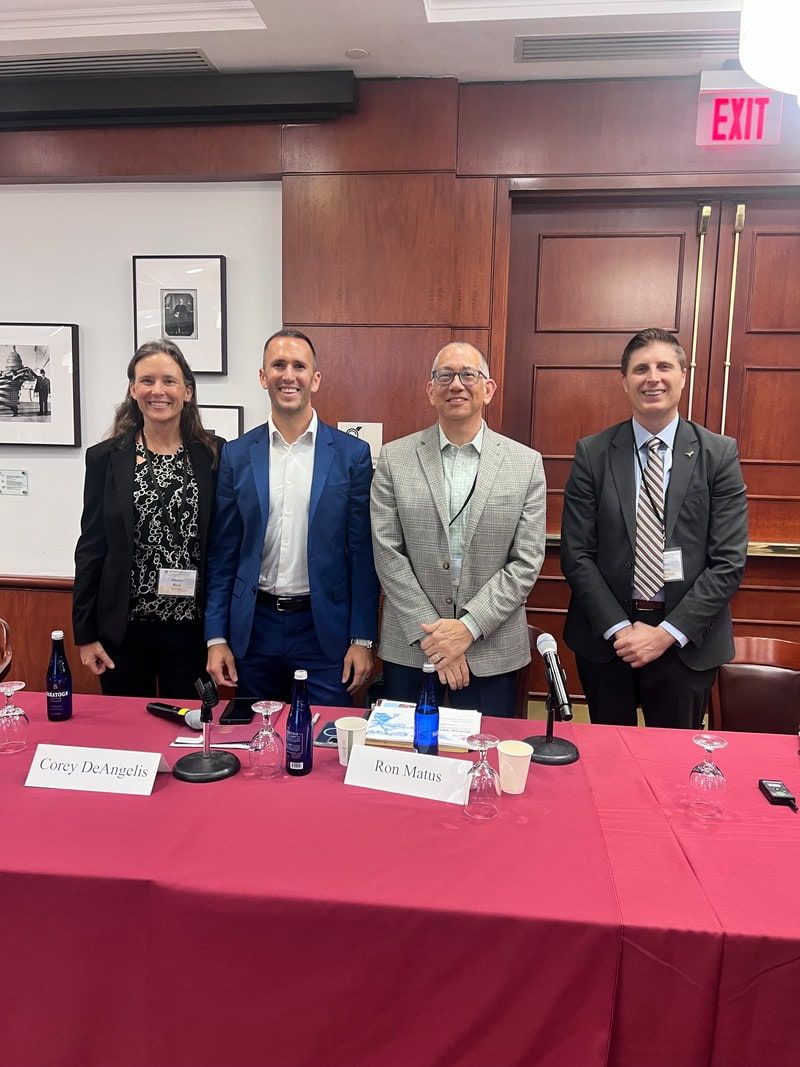 Four conference panelists standing behind a table with a red tablecloth, microphones and name cards.