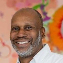 Jimmy Glover, smiling, with a gray beard and a white shirt, in front of a colorful mural.