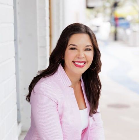 Leanndra Yates, smiling, with long dark hair and a light-pink blazer, seated on a step on a city sidewalk.