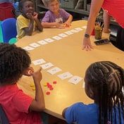 Children around a table laying out number cards while a teacher leans in to help.