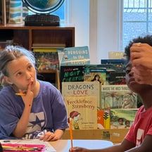 A girl in a star T-shirt holds a pencil to her chin at a table, with picture books on a shelf behind her.