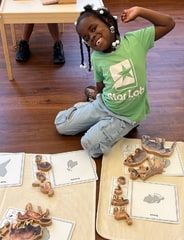 A girl in a green Star Lab T-shirt poses on the floor beside wooden animal puzzle pieces laid on picture cards.