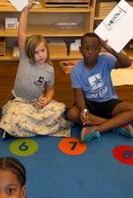 Children sitting on a rug printed with numbered circles, holding up small whiteboards.