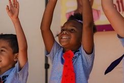 Children in light-blue shirts raise their arms; one wears a red paper tie.