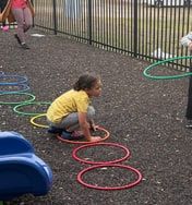 A child crouches beside colored hoops laid on an outdoor play area by a black fence.