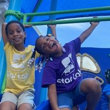 Two children in Star Lab T-shirts on a blue playground slide, one laughing with her mouth open.