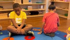 Children sitting cross-legged on a rug with numbered circles in a classroom with wooden shelves.