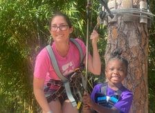 A smiling woman in a climbing harness beside a child in a harness next to a tree trunk.