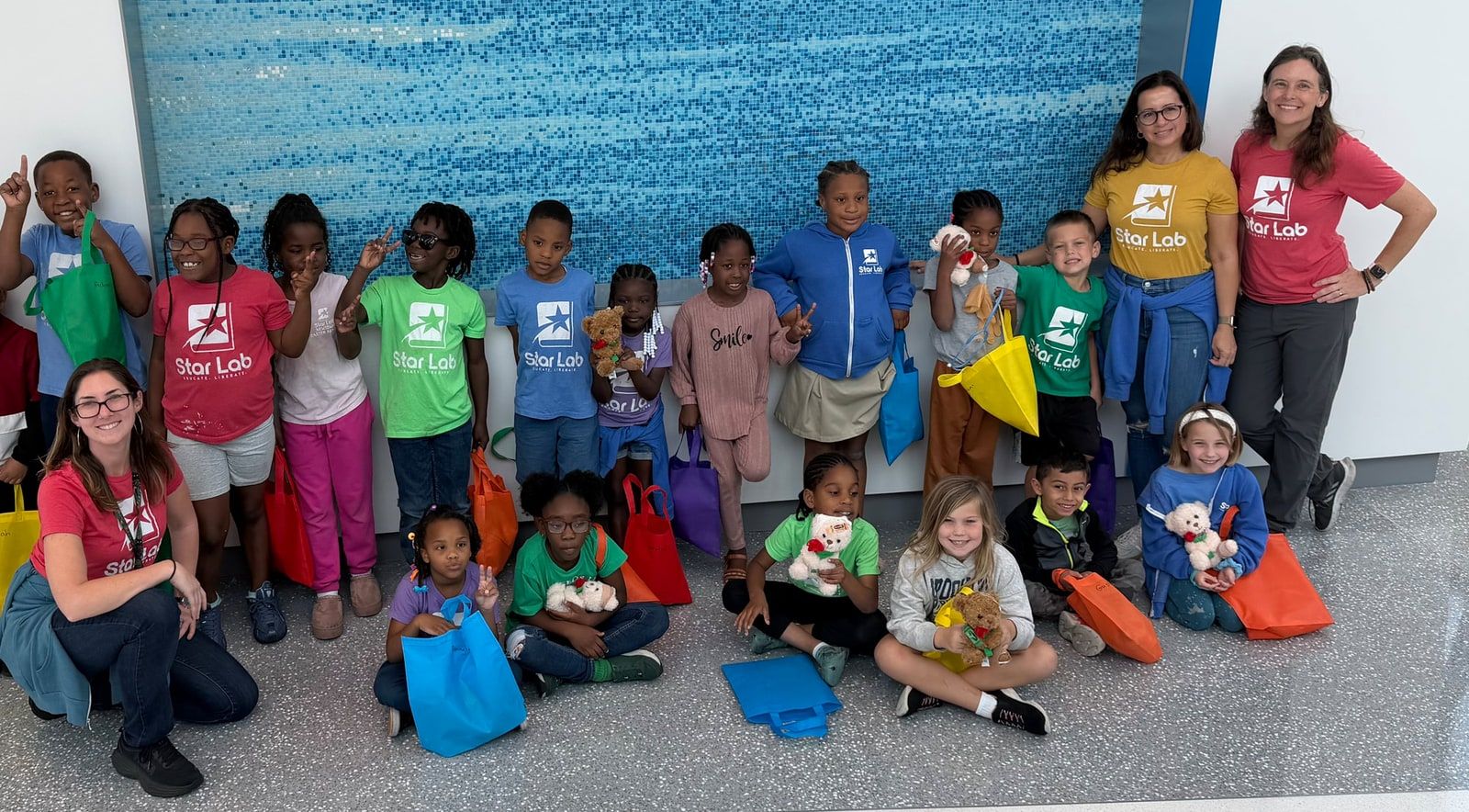 About twenty children and four adults, many in colourful Star Lab T-shirts, pose in front of a blue mosaic wall holding tote bags and stuffed animals.