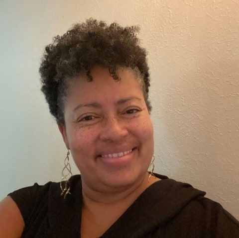 Mydahlia Glover, smiling, with short curly dark hair, a black top and drop earrings.