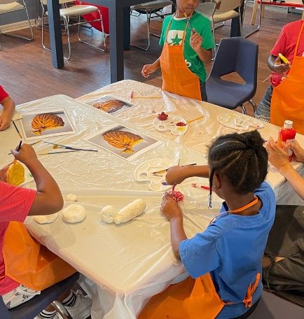 Children in orange aprons paint clay and pictures at a table covered with a plastic cloth.