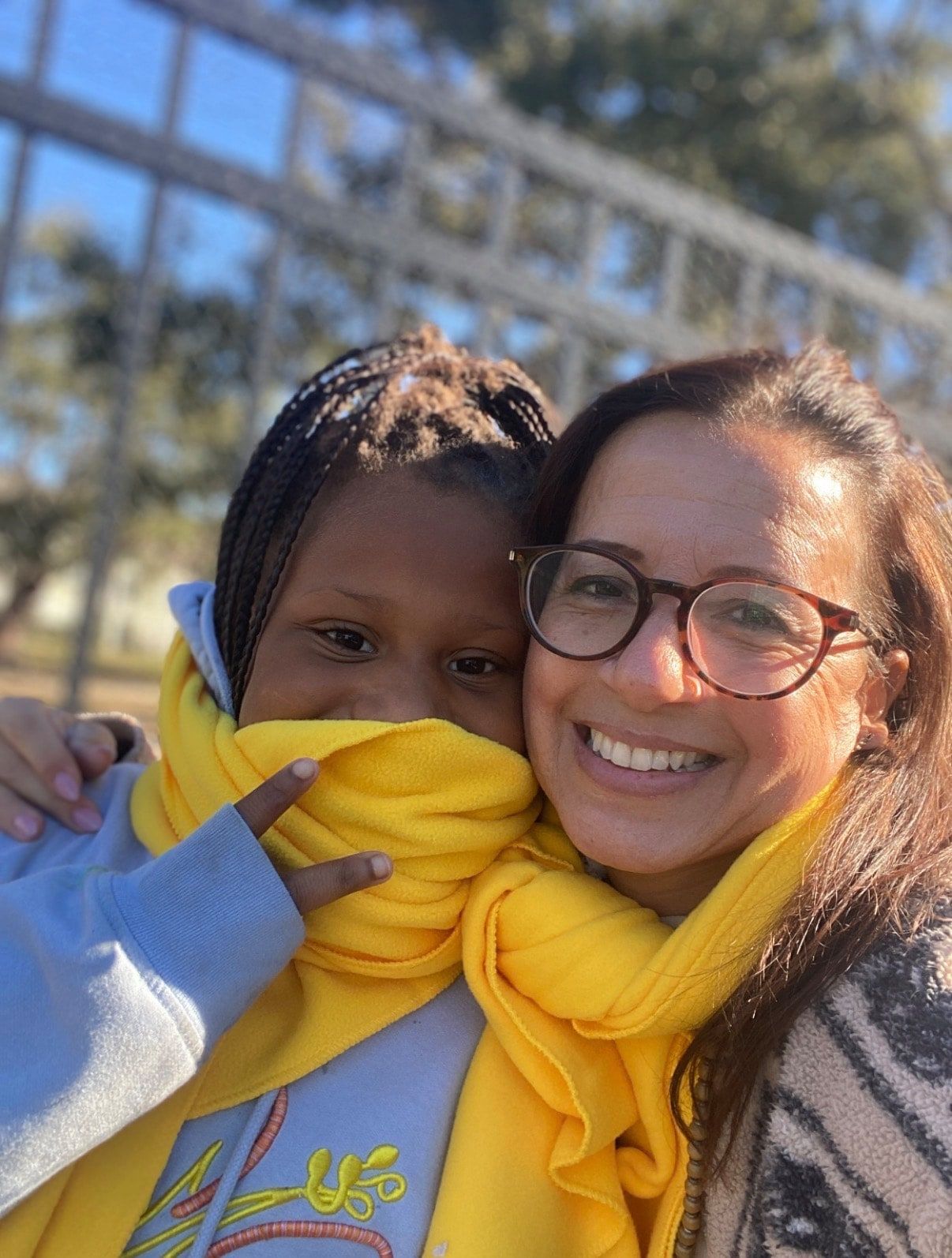 A smiling girl with braids and a yellow scarf pulled up over her mouth flashes a peace sign, cheek to cheek with a smiling woman in glasses, outdoors in front of a metal fence.