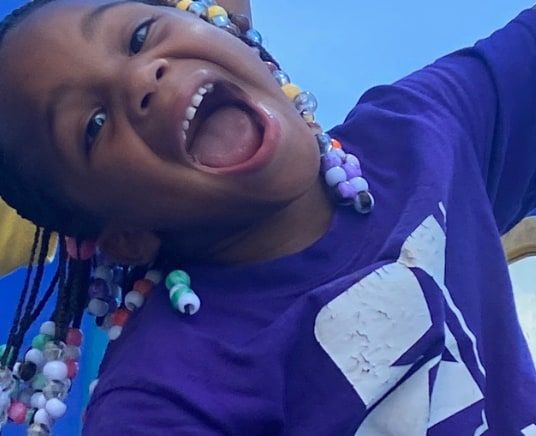 A laughing child with beaded braids in a purple Star Lab T-shirt on the playground.