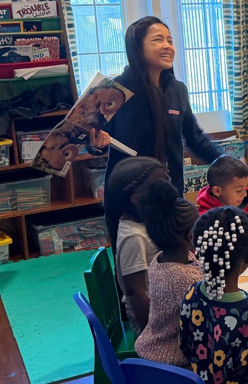 A smiling teacher holds up a picture book while children sit around a table.