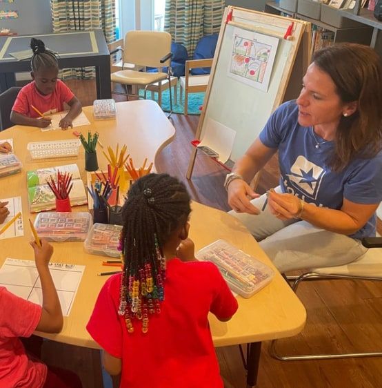 A teacher sits at a round classroom table with children who are writing and drawing, with cups of coloured pencils and supply trays on the table.