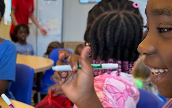 A smiling girl in class holding a green marker, with classmates working behind her.