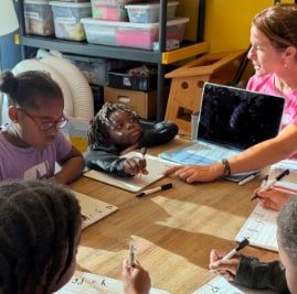 A woman in a pink shirt works with a laptop at a classroom table as children sit around the table writing on worksheets.