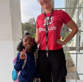 A woman in a red Star Lab T-shirt stands with her hand on her hip beside a smiling girl in a navy jacket who makes a peace sign.