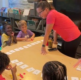 A woman in a pink shirt leans over a table of word cards while several children sit around the table.