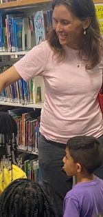 A smiling woman in a pink T-shirt points toward library shelves with children gathered in front of her.