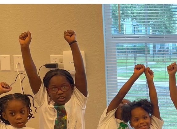 A group of children in white T-shirts with painted paper ties raising their fists in a classroom by a window.