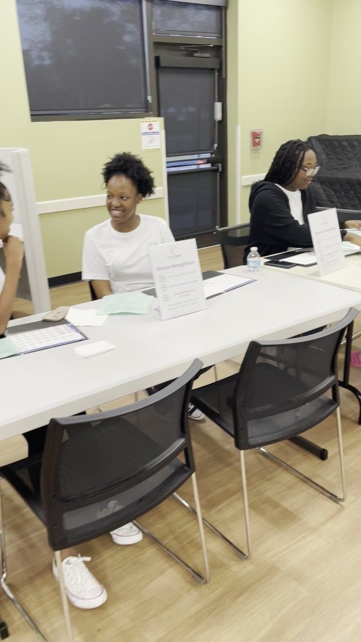 Two teenagers sit behind a long white table set with clipboards and table-tent signs in a community room; one smiles toward a person at the left edge of the frame.