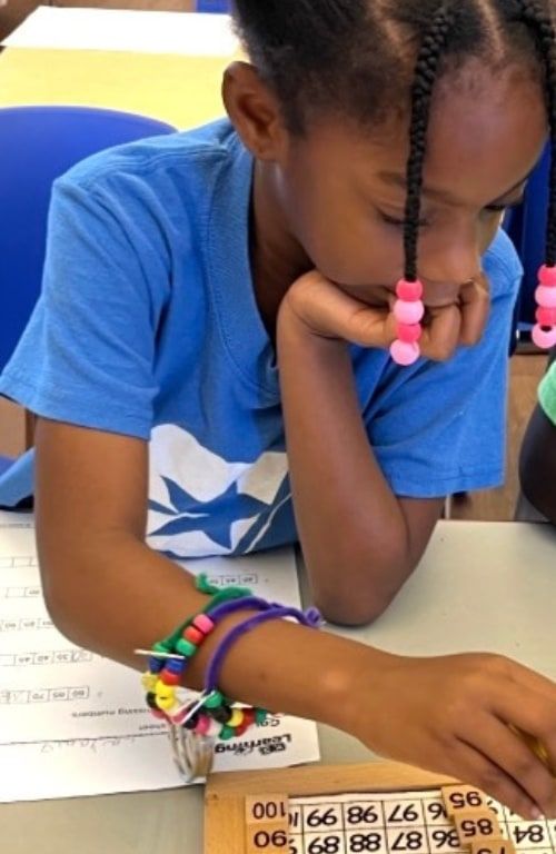 Two girls at a classroom table working on number worksheets; one rests her chin on her hand beside a wooden hundred-board puzzle.