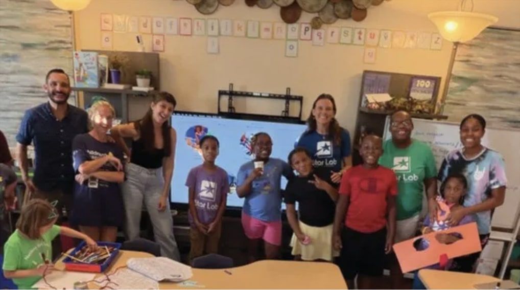 Group photo of teachers and children, several in Star Lab T-shirts, in a classroom with an alphabet banner and a large screen.